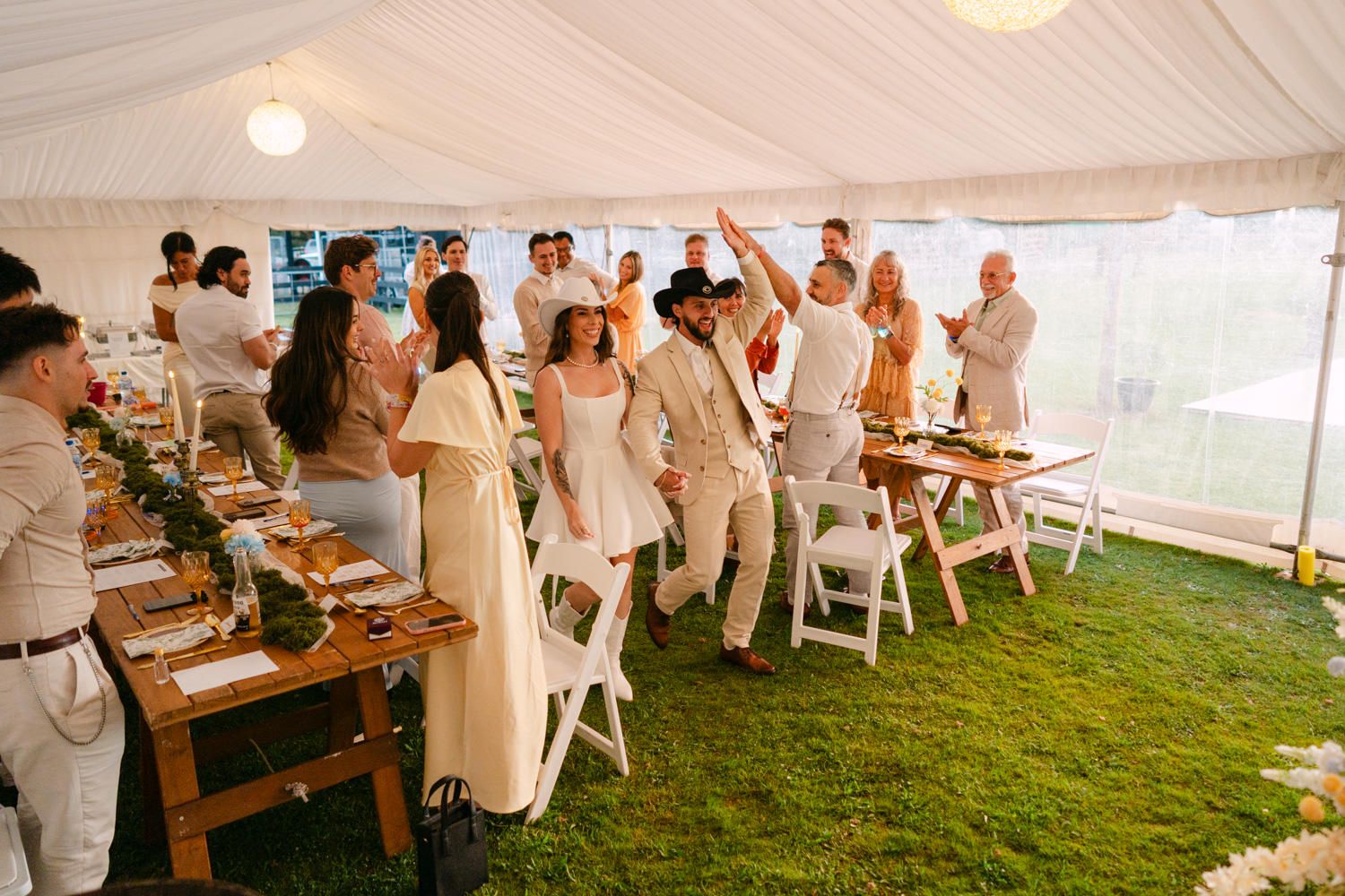 Guests celebrate under a white tent at an outdoor wedding reception with long wooden tables on green grass.