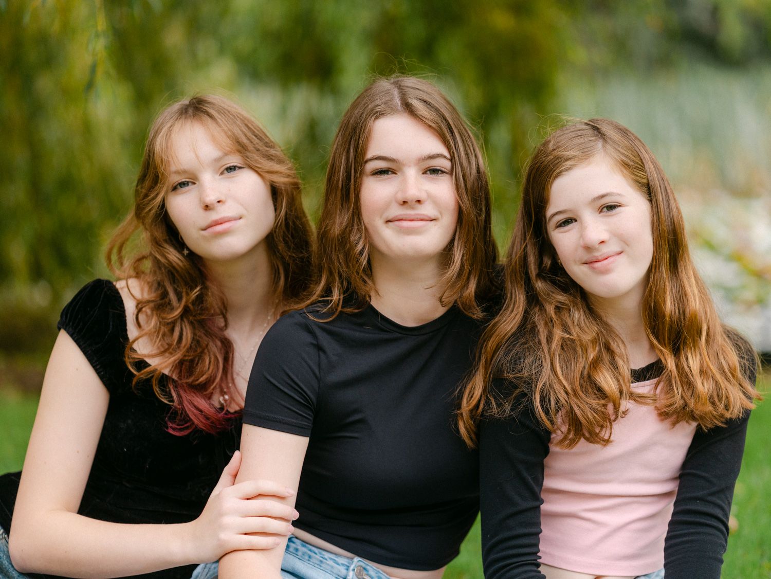 Three teenage girls with auburn hair pose together outdoors, smiling in casual attire against a green tree backdrop.