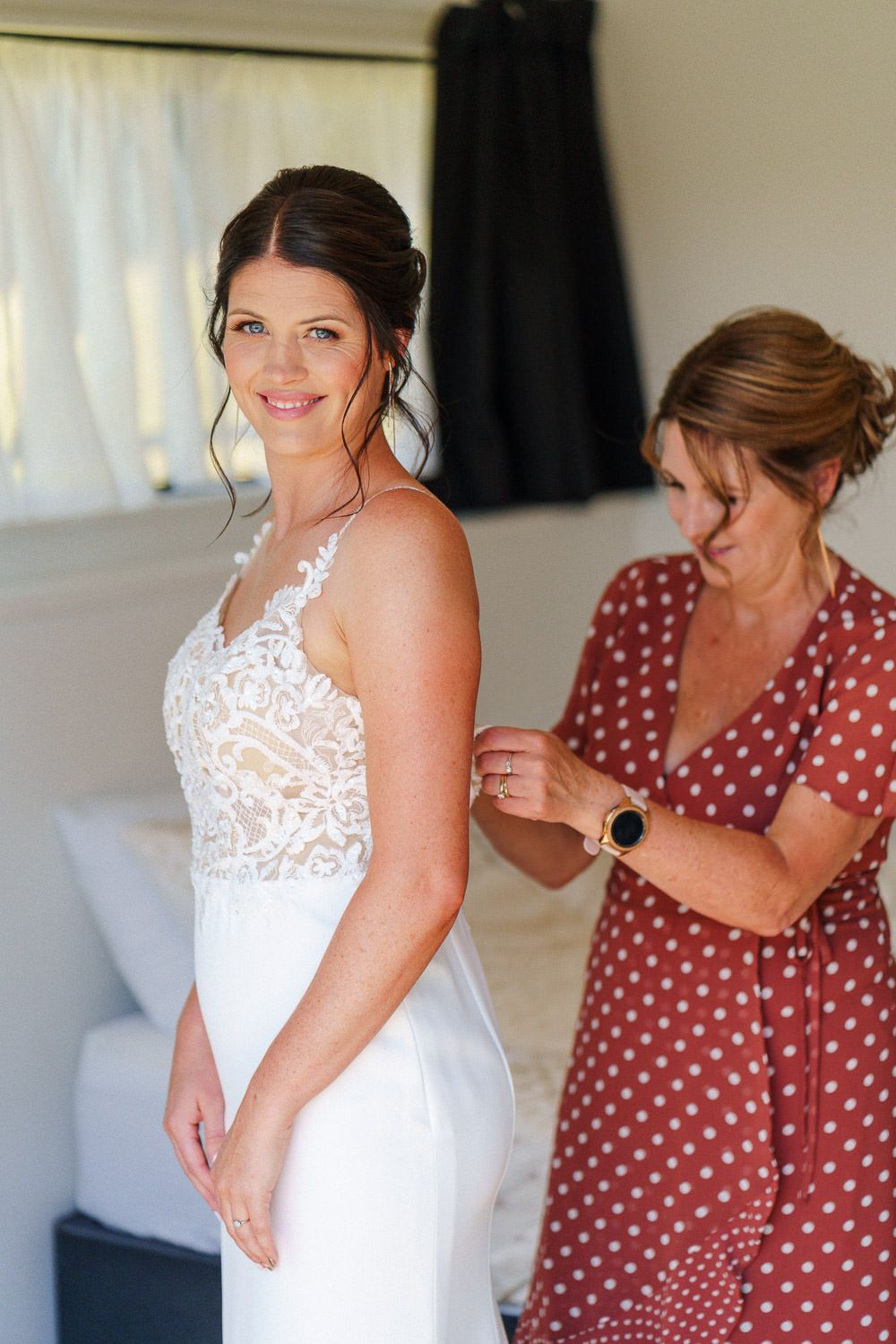 Bride in white lace wedding dress smiles as woman in red polka dot dress fastens the back of her gown.
