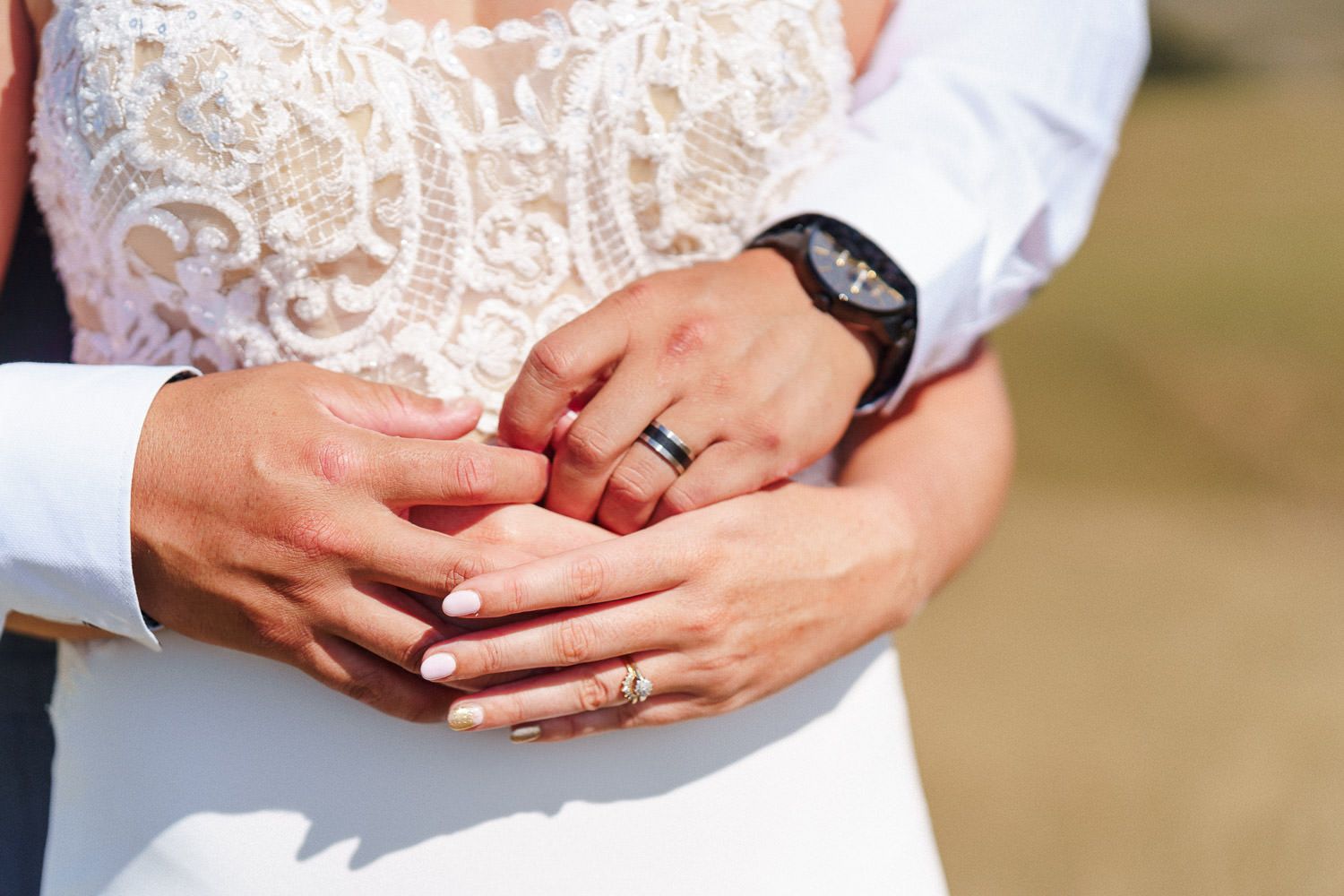Close-up of newlywed couple holding hands showing wedding rings, bride in lace dress, groom in suit.
