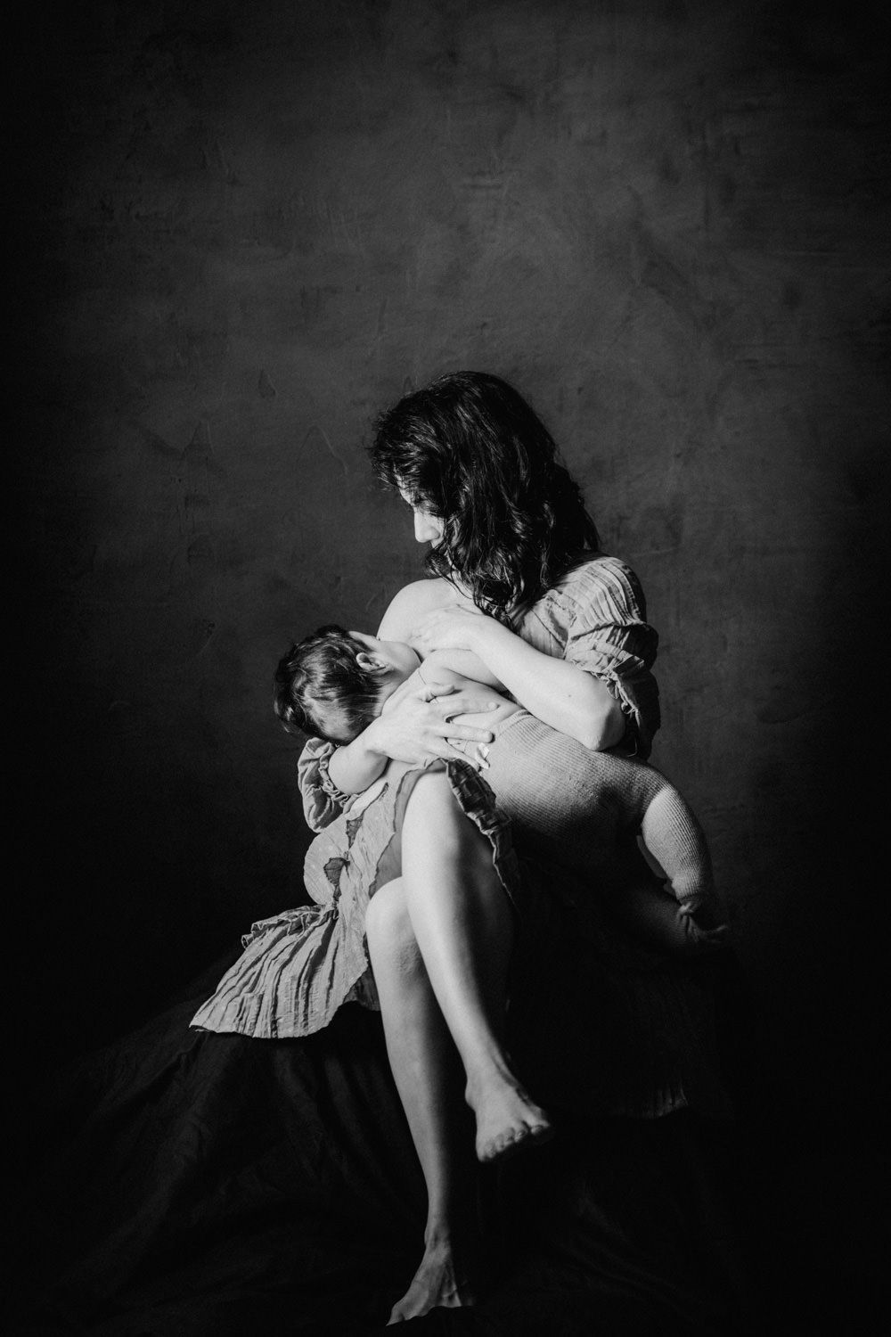 Black and white photo of a mother tenderly holding her baby while seated against a dark studio background.
