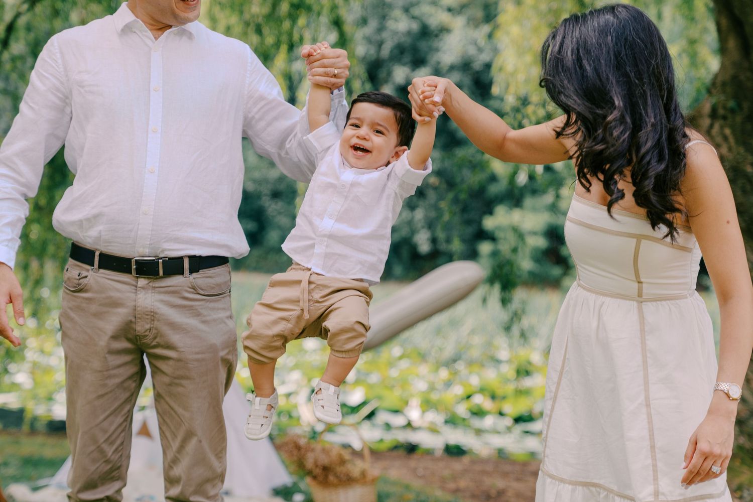 Parents joyfully swing their laughing toddler boy between them in a sunny green park during a family photo session.