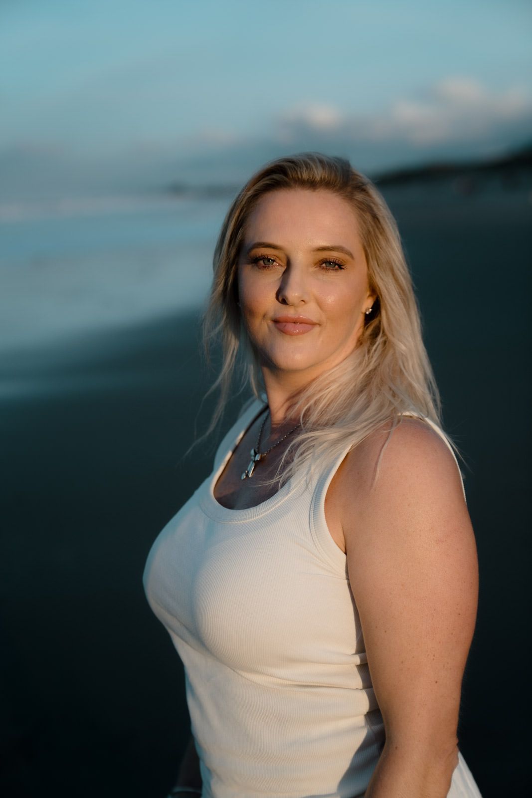 Blonde woman in white tank top posing on beach at sunset with ocean waves in background.