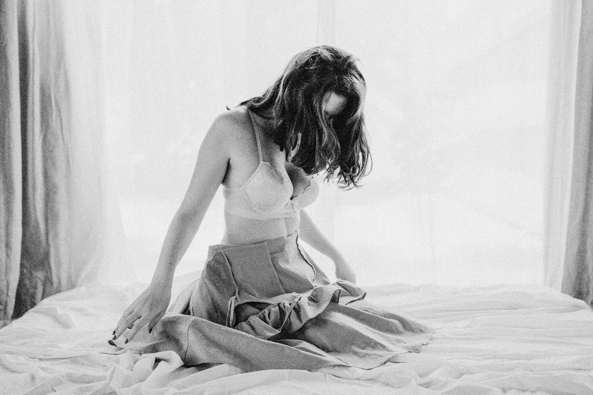 Black and white photo of a woman in a skirt sitting on a bed near sheer curtains in soft natural light.