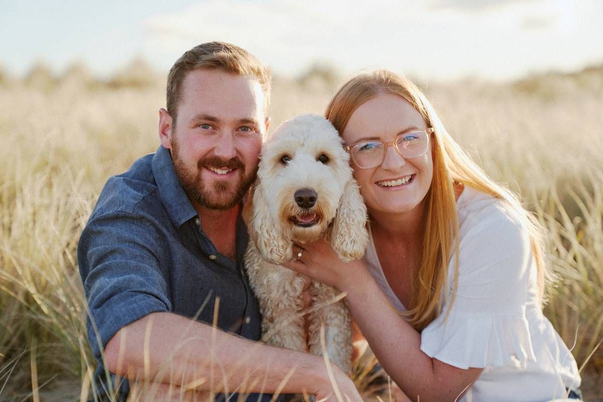 Smiling couple posing outdoors in grassy dunes with a fluffy white Goldendoodle dog on a sunny day.