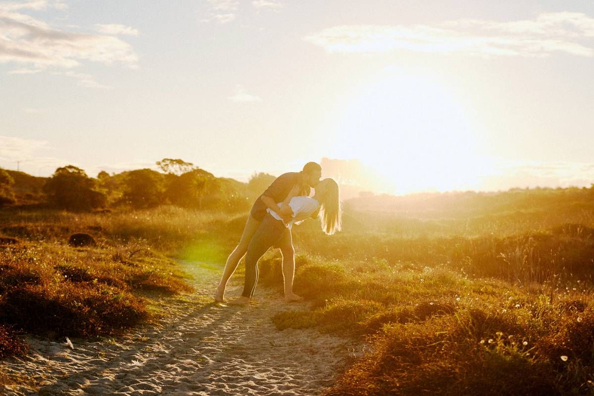 Couple sharing a romantic kiss on a sandy path at golden sunset in a scenic outdoor landscape.