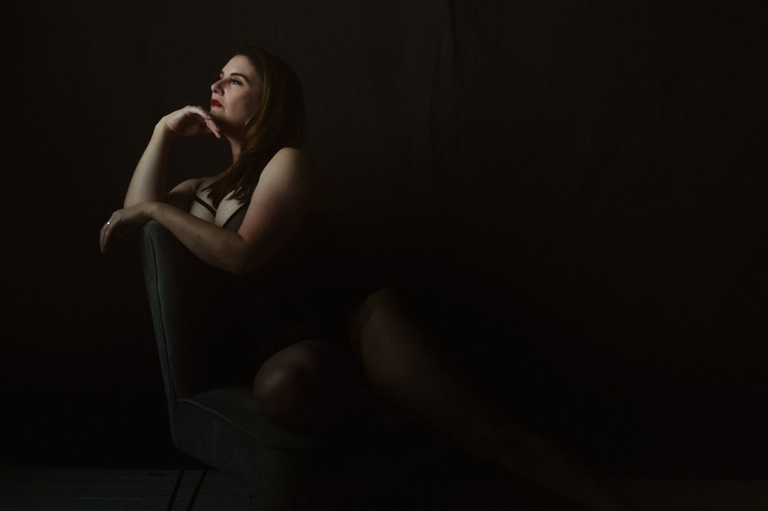 A pensive young woman sits alone in dramatic low-key lighting, gazing upward in a contemplative pose.