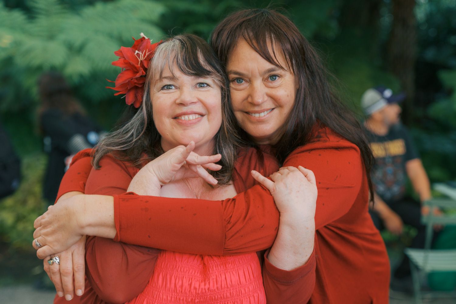 Two women in red outfits smiling and embracing outdoors, one wearing a red flower in her hair.