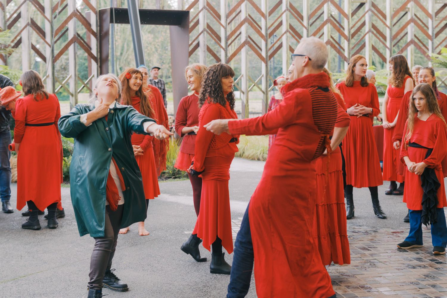 Women in red and green dresses dancing together outdoors near a decorative wooden building.