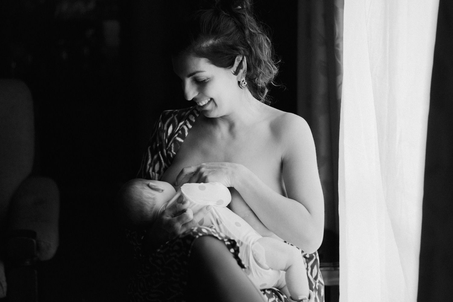 Black and white photo of a smiling mother breastfeeding her newborn baby near a window.