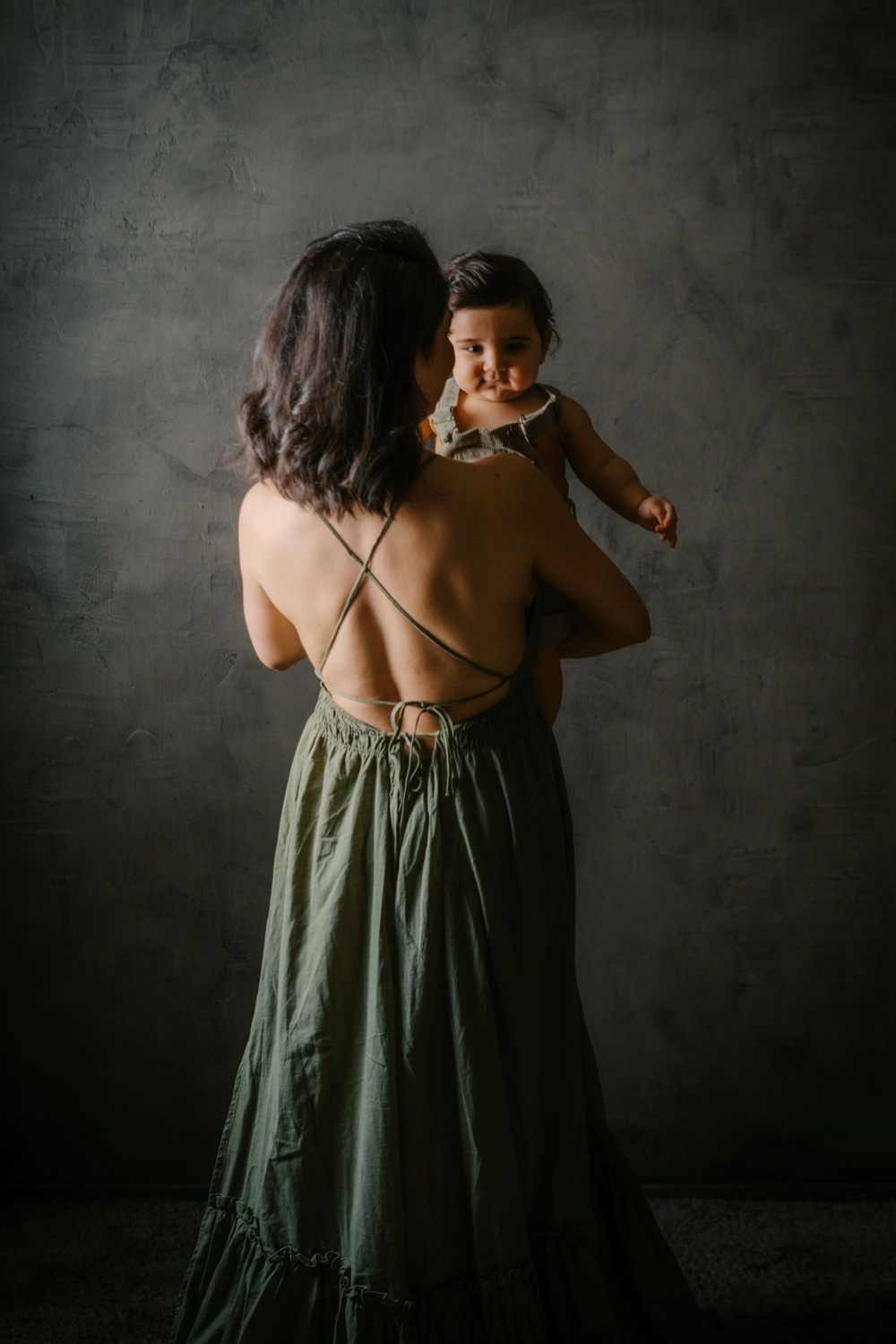 A woman in a backless green maxi dress holds a smiling baby against a dark gray studio backdrop.