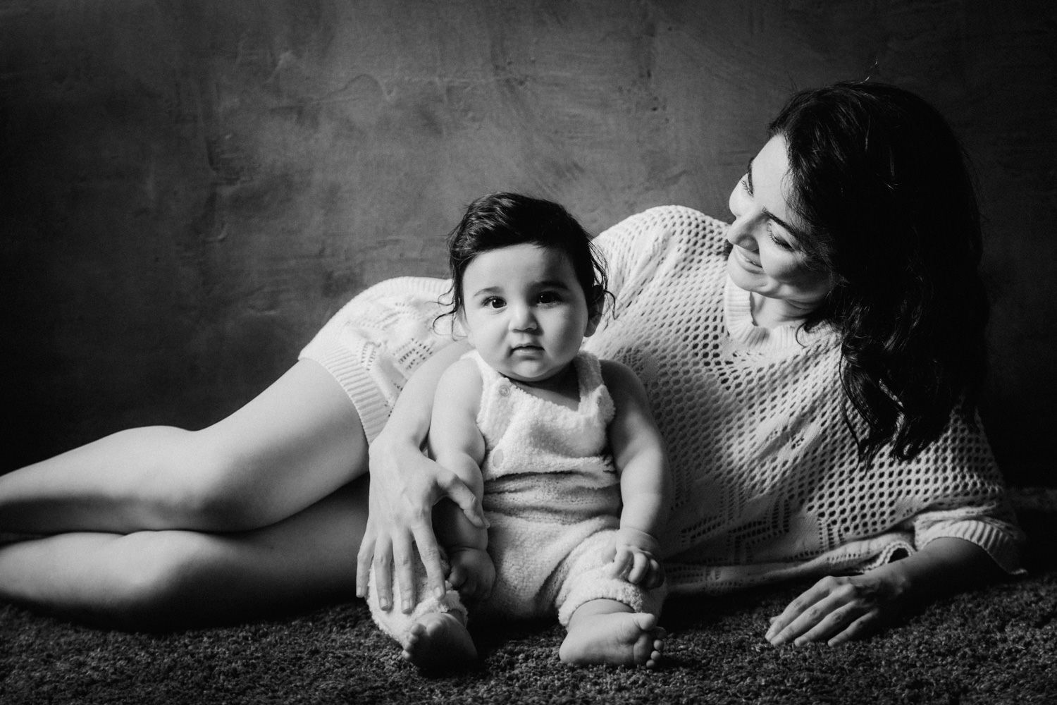 Black and white photo of a mother lying down beside her infant baby on a textured carpet in a studio.