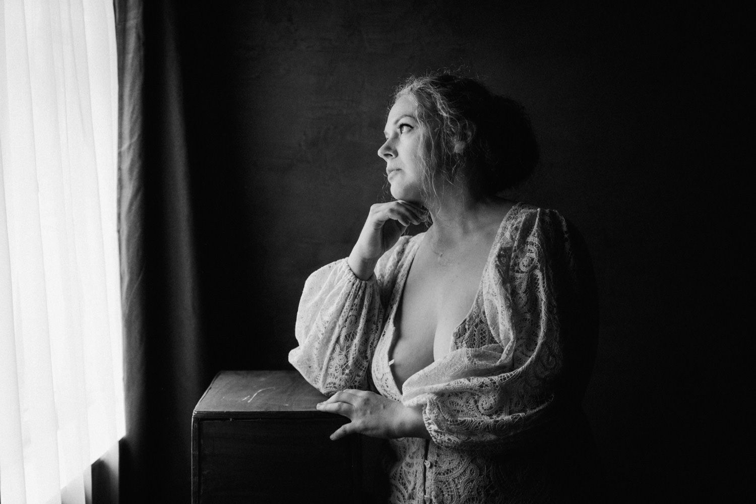 Black and white portrait of a woman in lace robe gazing thoughtfully out a window in soft natural light.