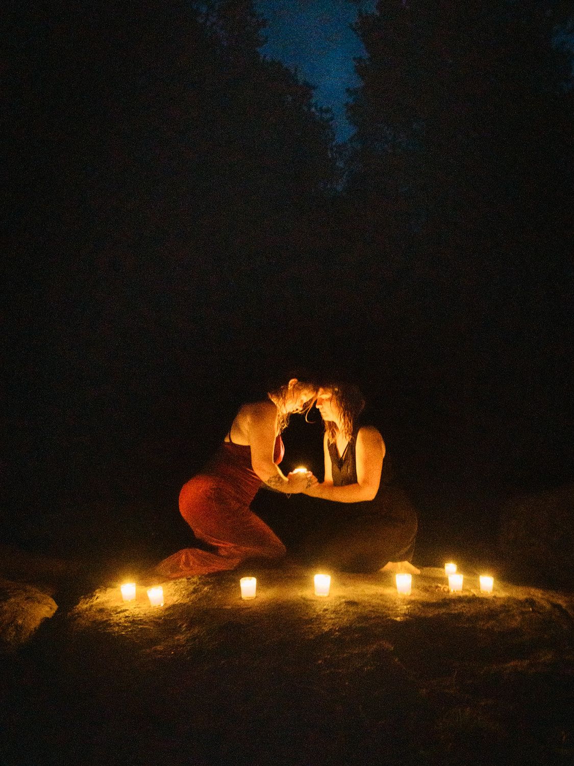 Two people share a romantic moment surrounded by glowing candles outdoors at night under a dark starry sky.