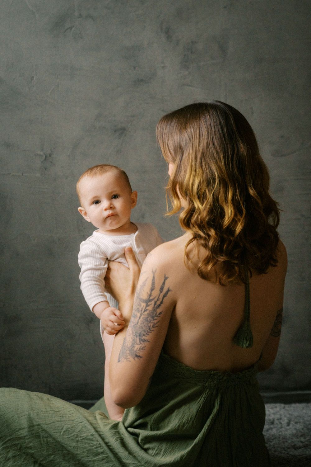 A tattooed mother holds her baby in a studio, wearing a green dress, with a gray textured wall background.