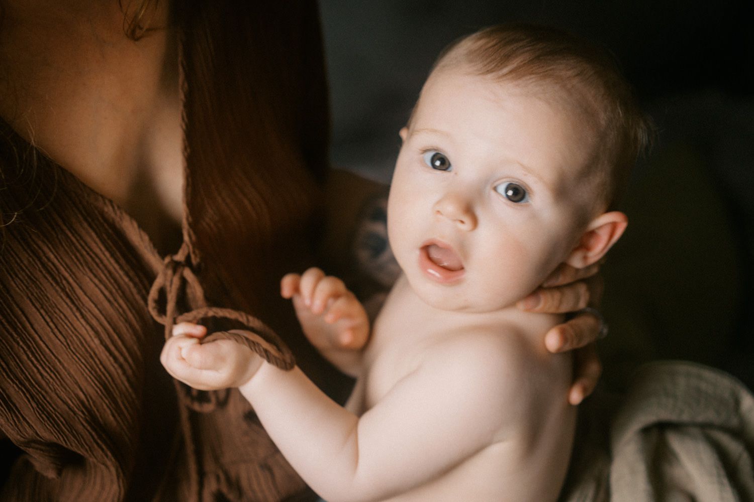 Adorable baby with blue eyes held by mother in brown dress, looking curiously at camera in warm light.