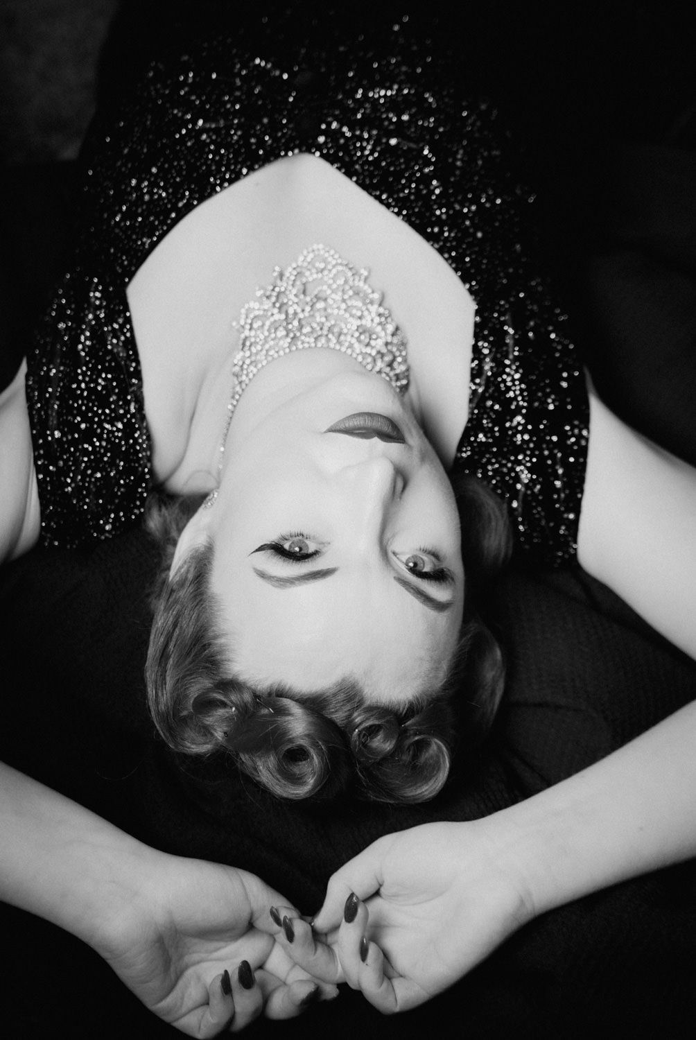 Black and white photo of a woman lying down wearing a sparkly dress and crystal tiara, looking up at camera.