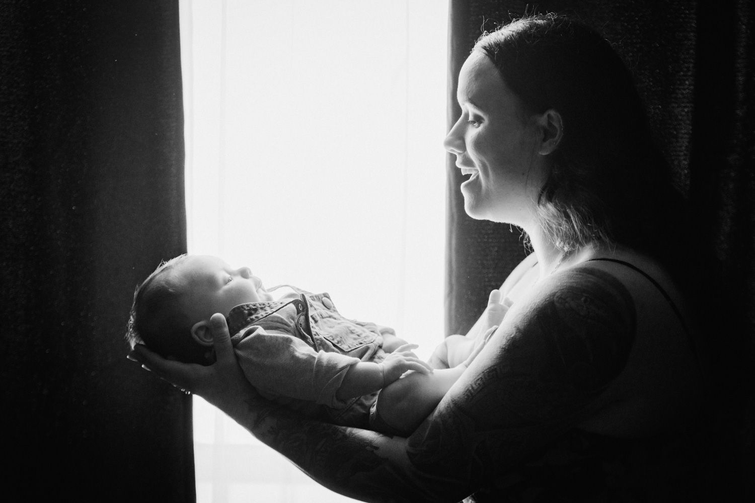 Black and white photo of a mother cradling her newborn baby by a bright window, sharing a tender moment.