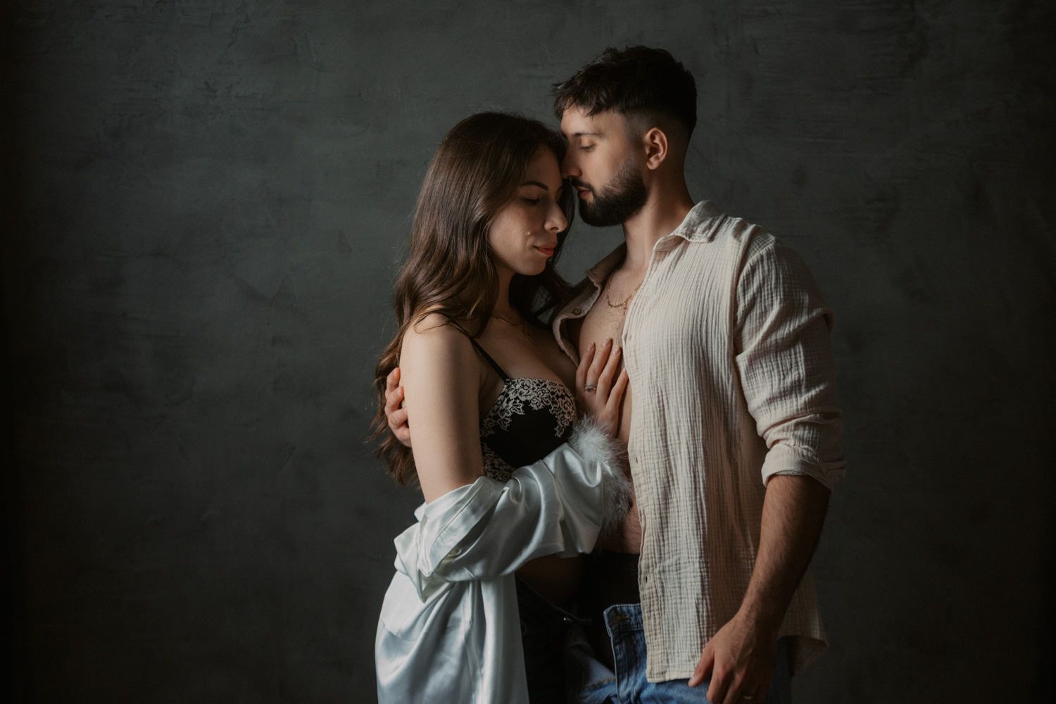 Romantic couple embracing intimately in a studio, woman in lace top and man in white shirt against dark background.