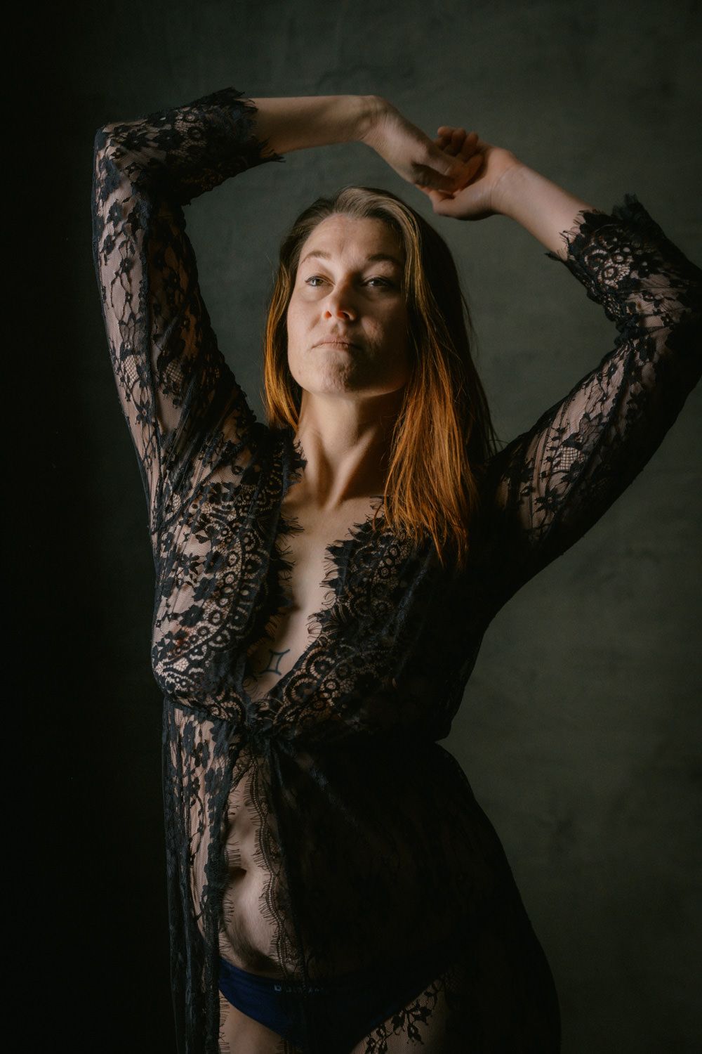 Woman in sheer black lace robe posing with arms raised against dark background.