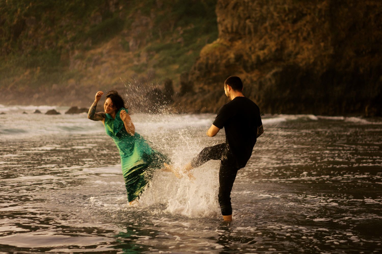 Couple splashing water playfully in ocean waves near a lush green cliff at golden hour.