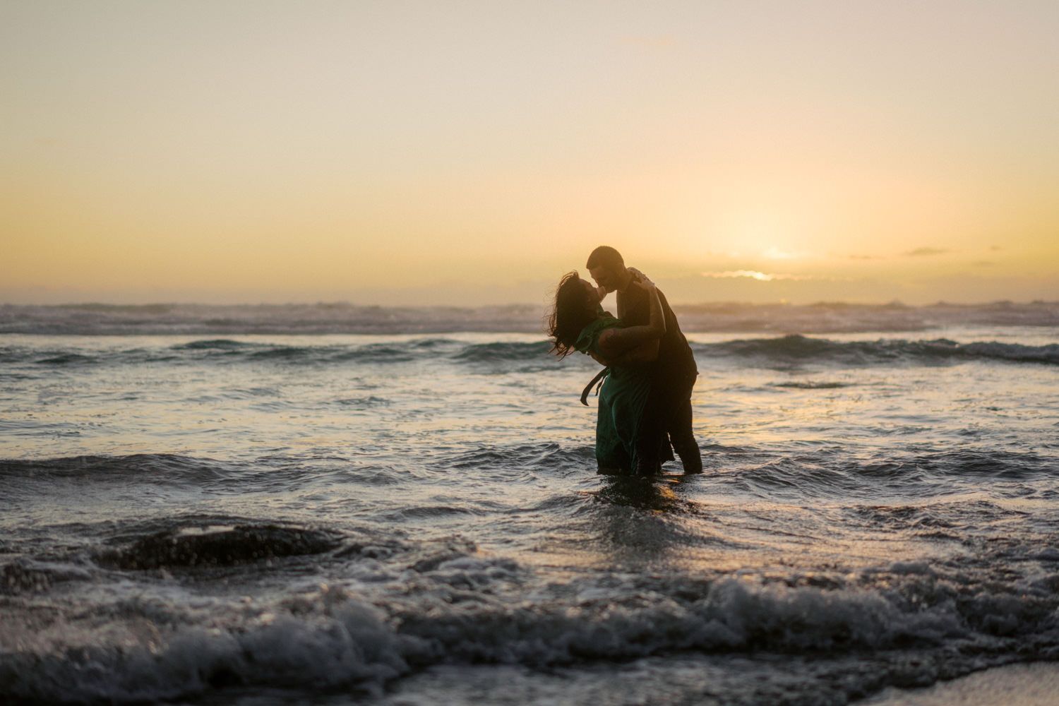 Couple silhouetted against golden sunset waves on beach, embracing romantically in the ocean surf.