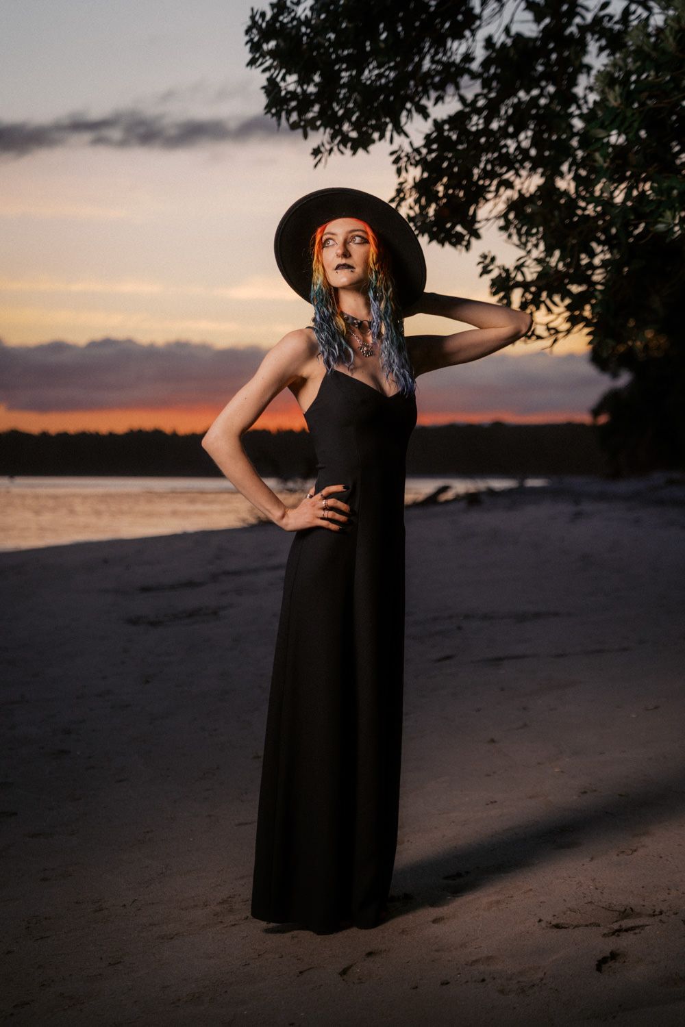 Woman in black gown and wide-brim hat poses on sandy beach at sunset with colorful braided hair.