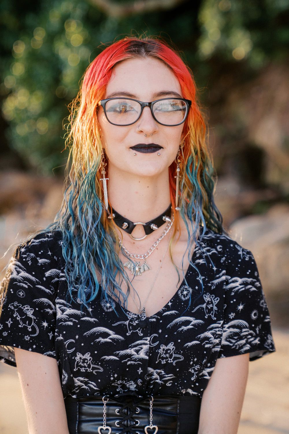 Young woman with rainbow-dyed hair, black lipstick, glasses, and gothic-style jewelry wearing a celestial print black t-shirt.