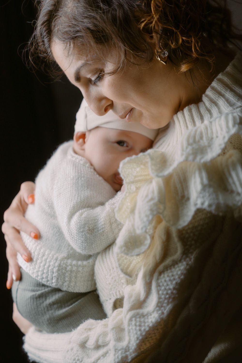 A curly-haired mother in a white knit sweater tenderly holds her newborn baby close to her chest.