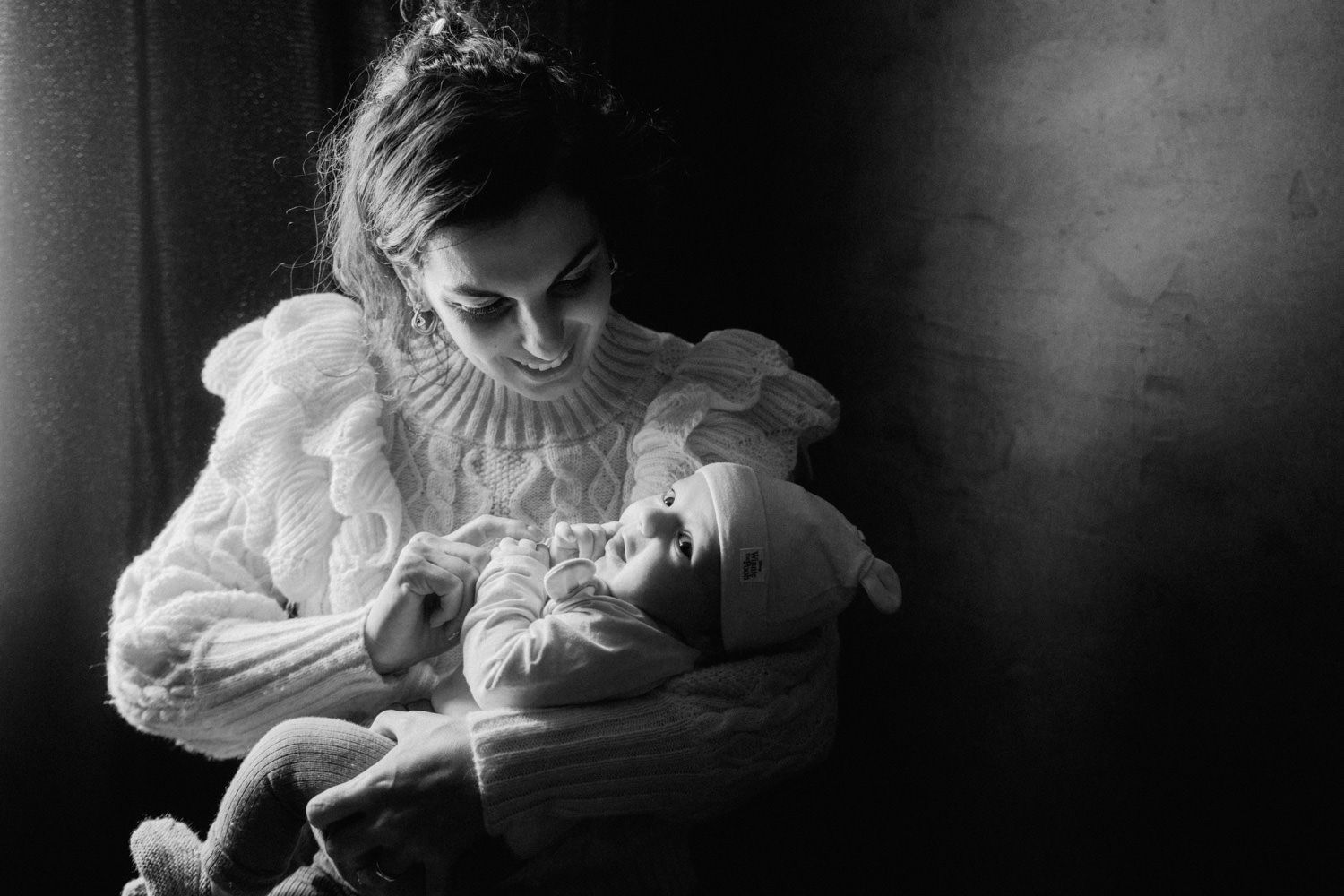 Black and white portrait of a mother smiling at her newborn baby in soft dramatic lighting.