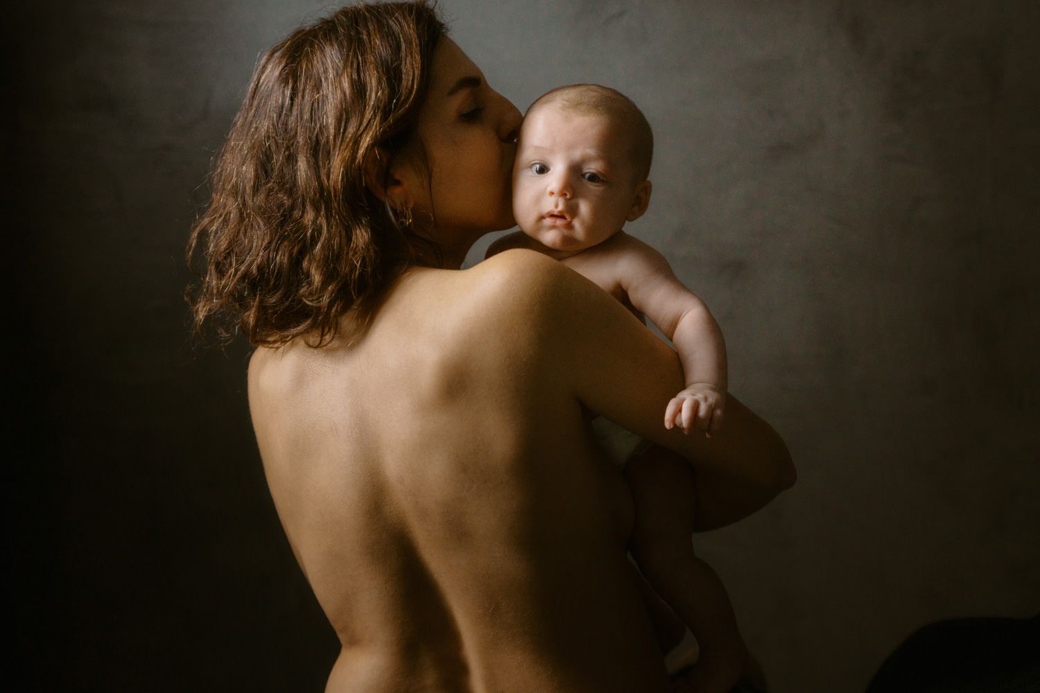A mother with bare back tenderly kisses her newborn baby against a dark studio background.