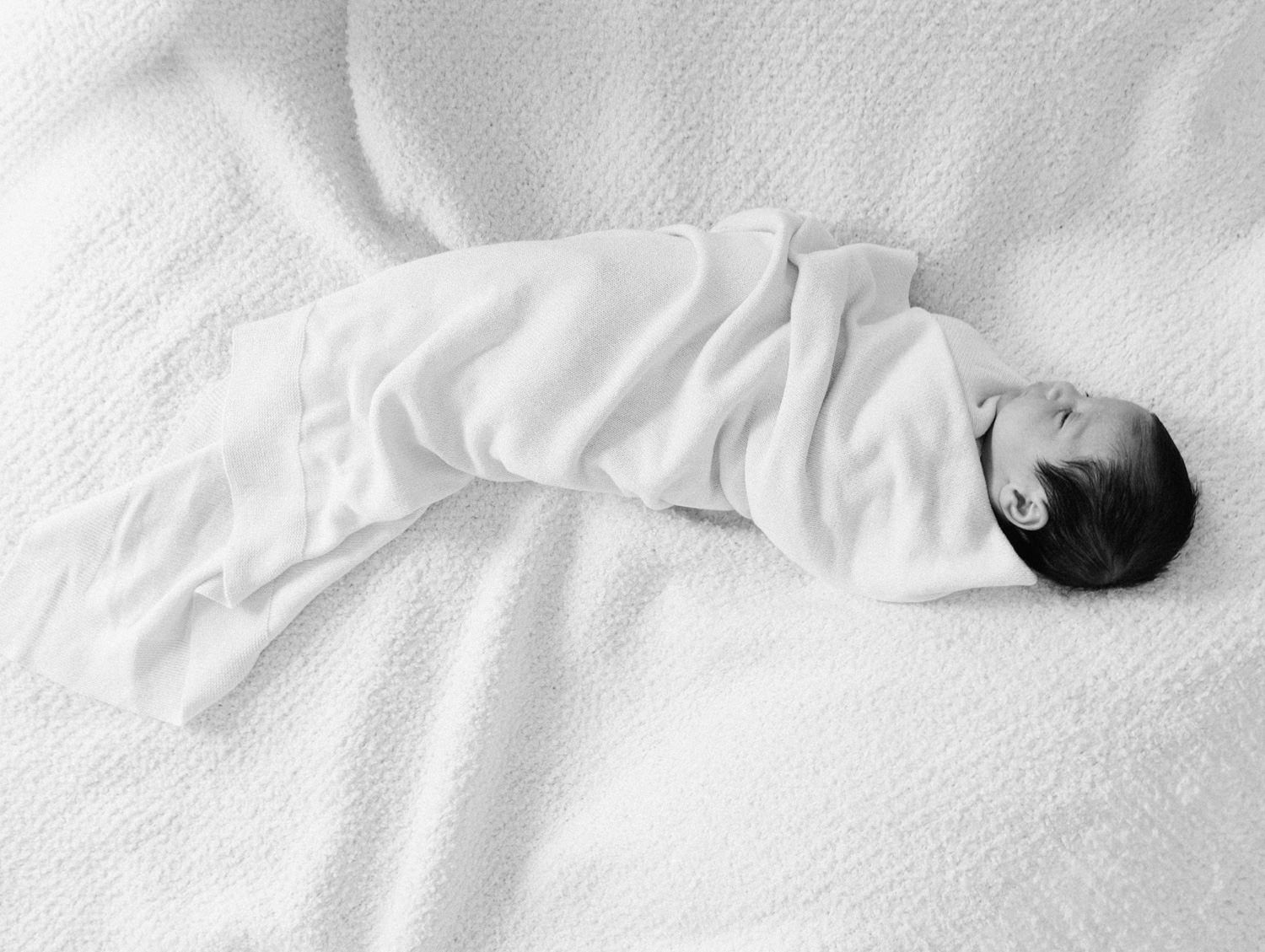 Black and white photo of a swaddled newborn baby lying on a white textured blanket.