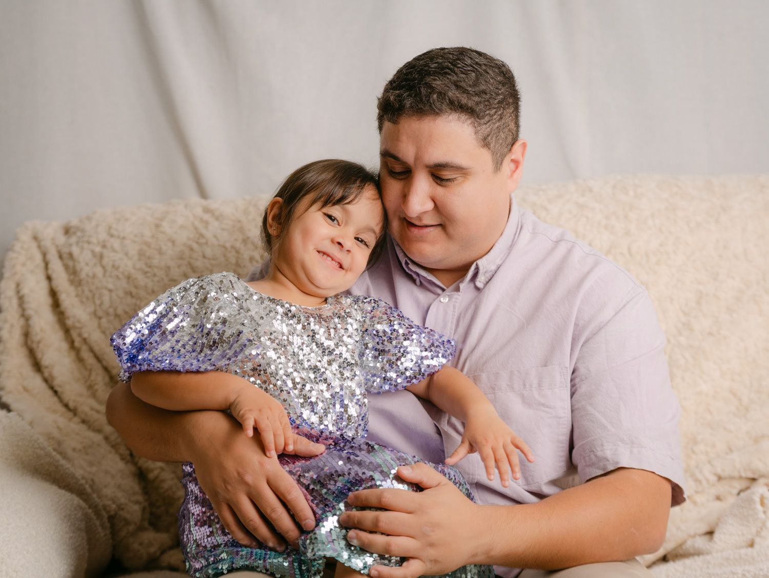 A father and young daughter in a sequin dress share a tender moment on a cozy couch in a studio portrait.