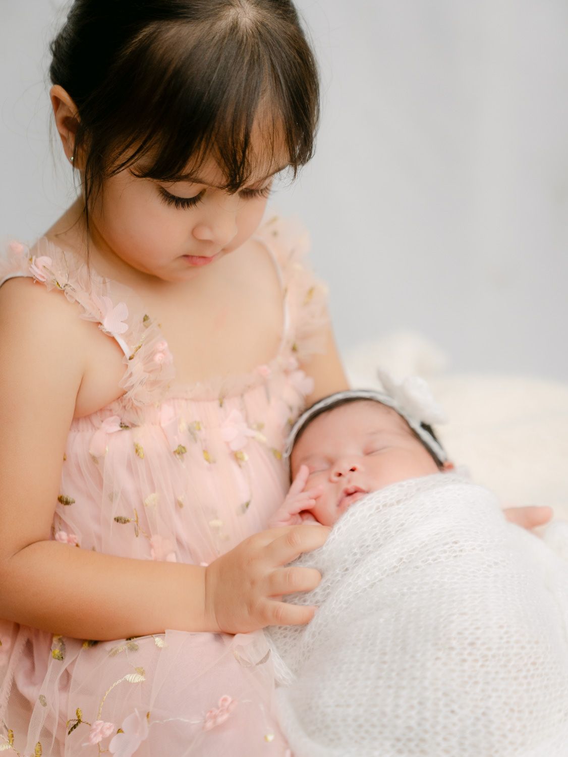 A young girl in a pink floral dress tenderly holds a sleeping newborn wrapped in a white swaddle.