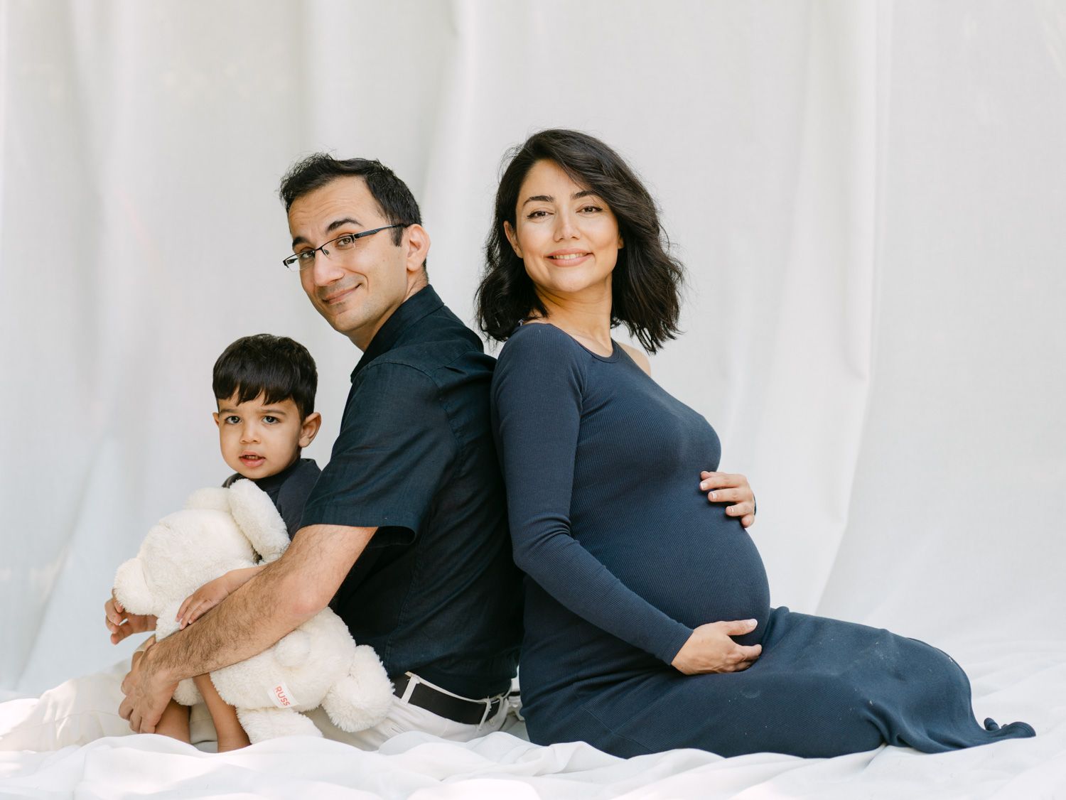 Family maternity photo with pregnant woman in navy dress, man in black shirt, and toddler holding teddy bear.