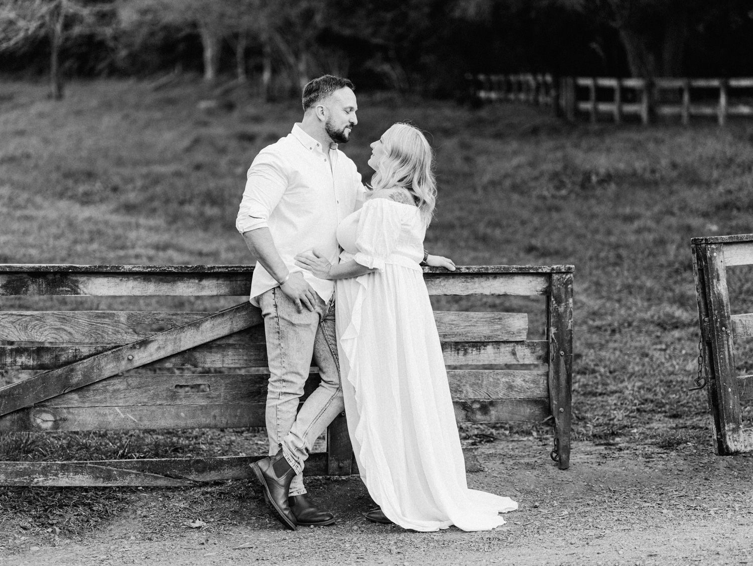 Black and white maternity photo of couple leaning against rustic fence in countryside setting.