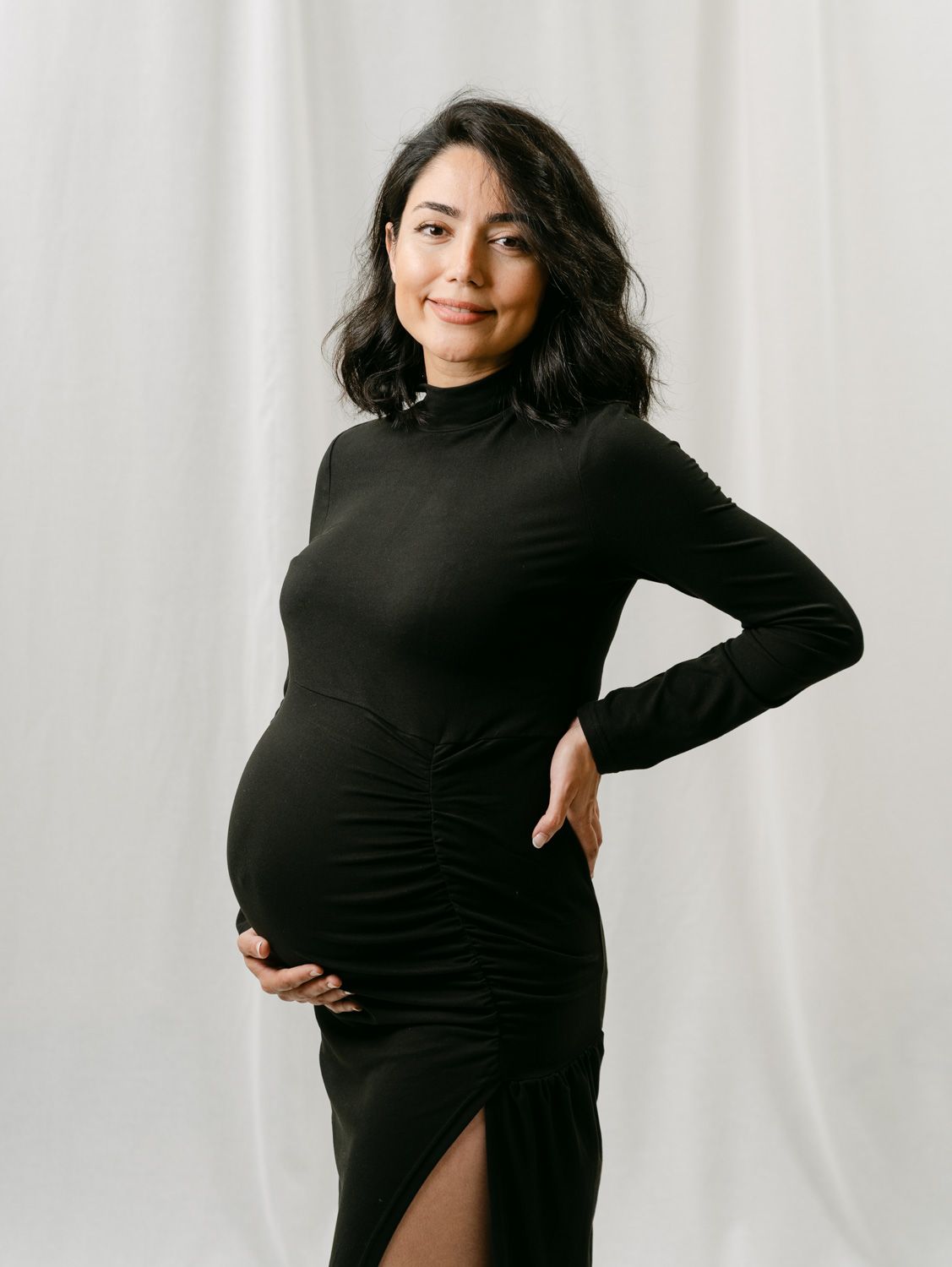 Pregnant woman in black long-sleeve ruched maternity dress posing gracefully against white backdrop.