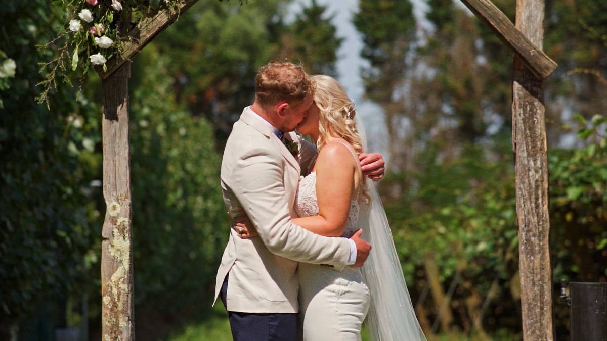 A bride and groom share their first kiss under a rustic wooden arch adorned with flowers at an outdoor wedding.