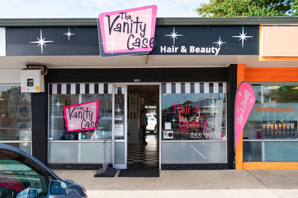 Exterior of The Vanity Case Hair & Beauty salon with black facade, striped awning, and pink signage.