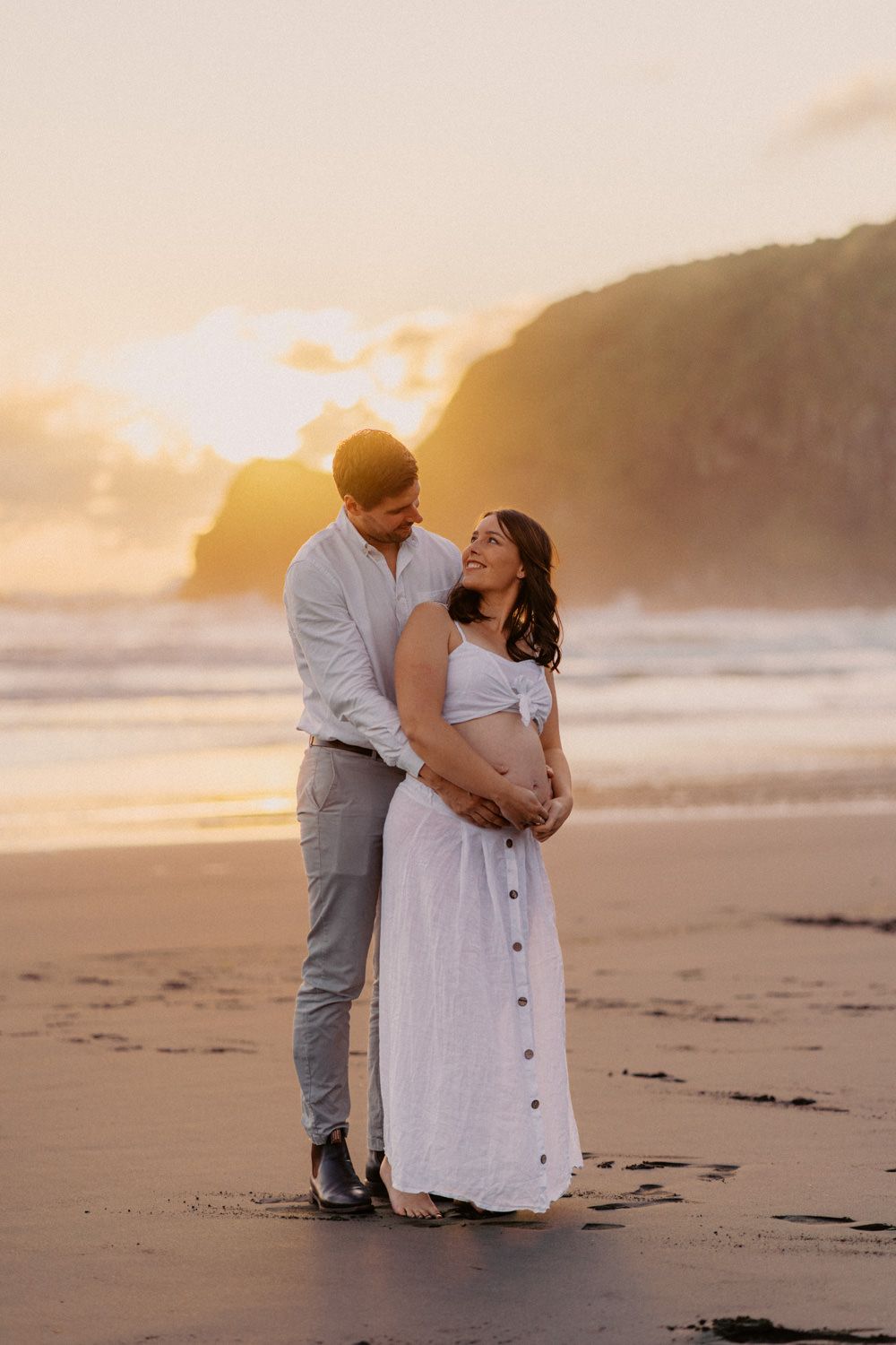 Couple embracing on beach at golden sunset, pregnant woman in white dress, man in white shirt, ocean and cliffs backdrop.