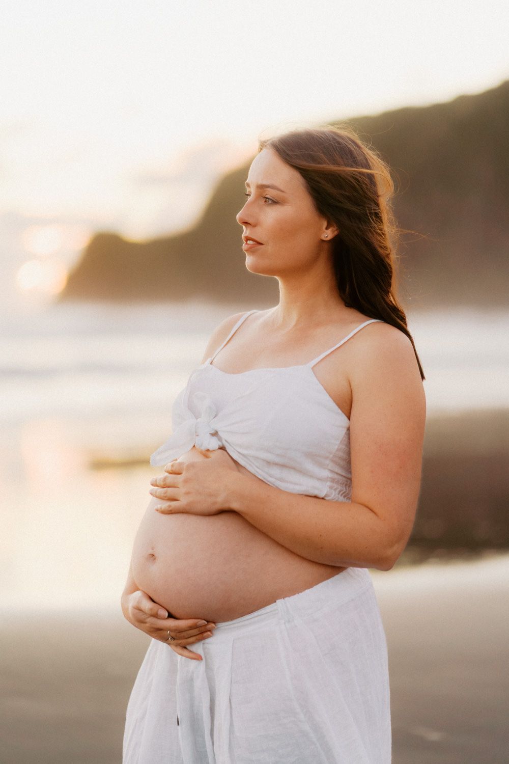 Pregnant woman in white dress cradling her baby bump on a beach at golden hour sunset.