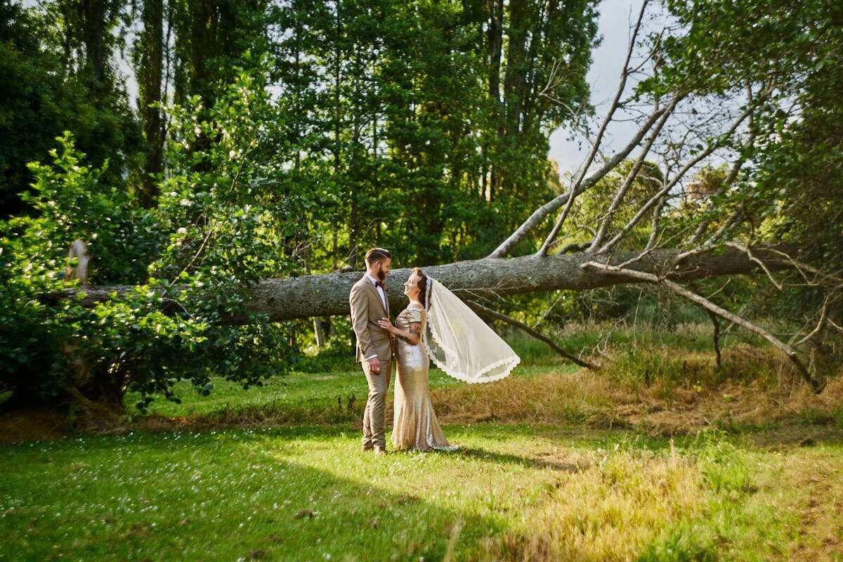 Bride and groom share a romantic moment in a lush green garden with a fallen tree backdrop.
