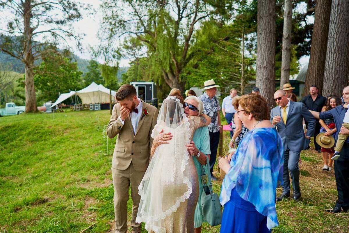 Emotional groom covering his face as bride walks toward him during outdoor wedding ceremony on green lawn.