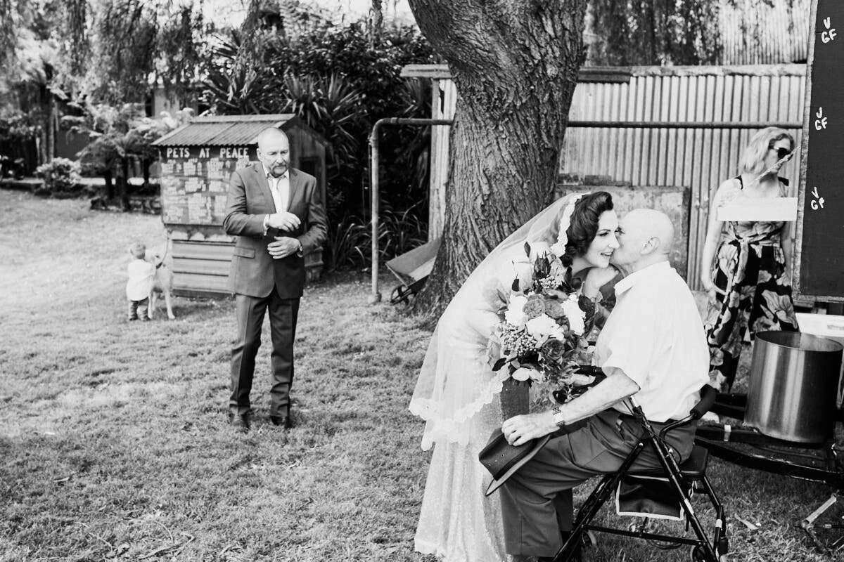 Black and white wedding photo of bride kissing elderly man in chair as groom watches outdoors near a large tree.
