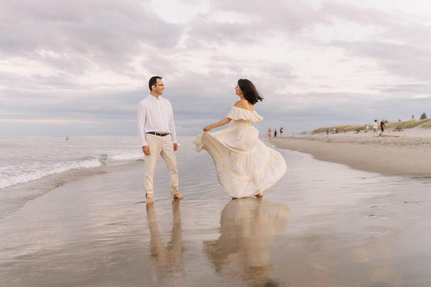 Couple in white outfits dancing on a beach at sunset with waves and cloudy sky.