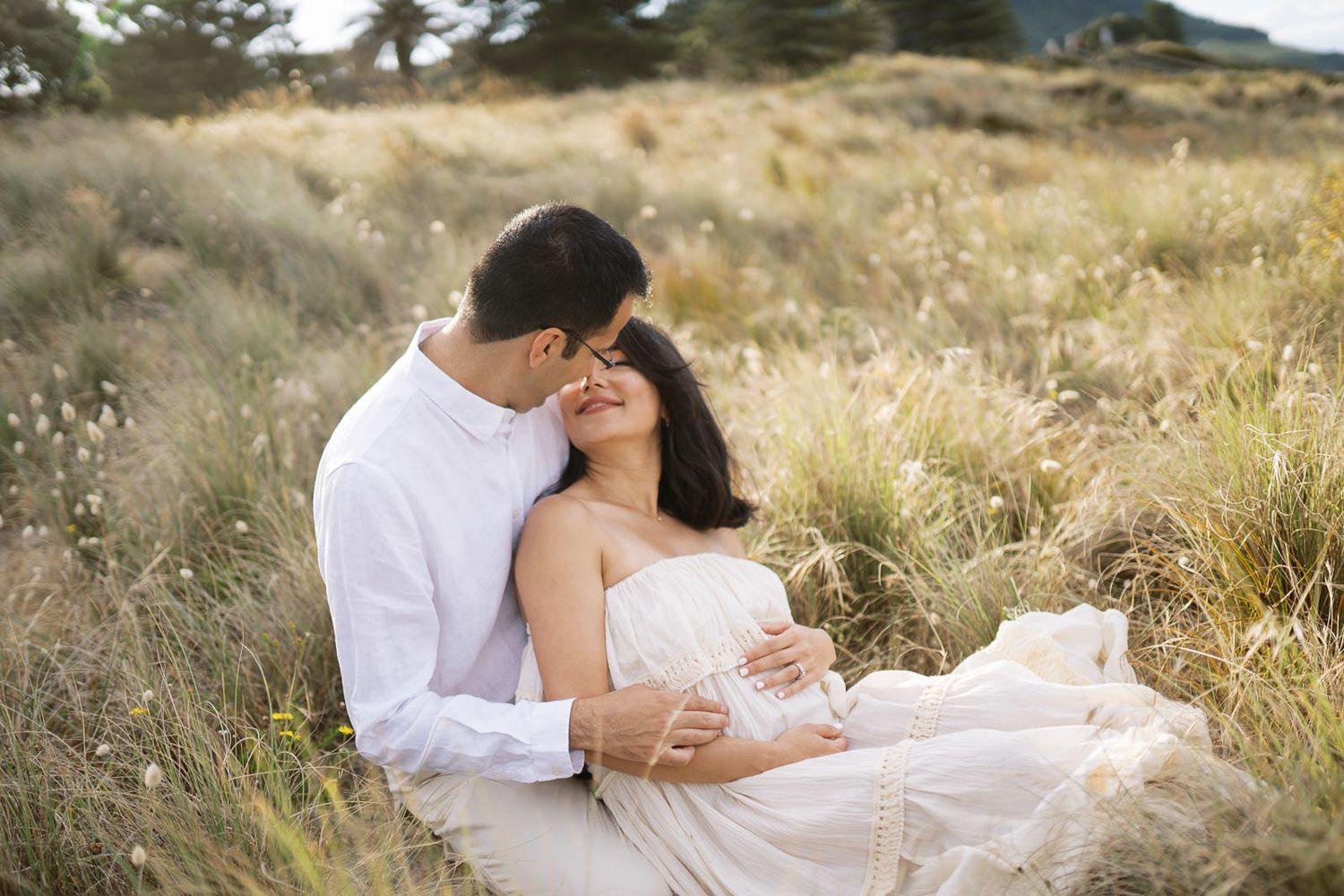 Romantic couple embracing in golden meadow, woman in white dress, man in white shirt, outdoor engagement photo.