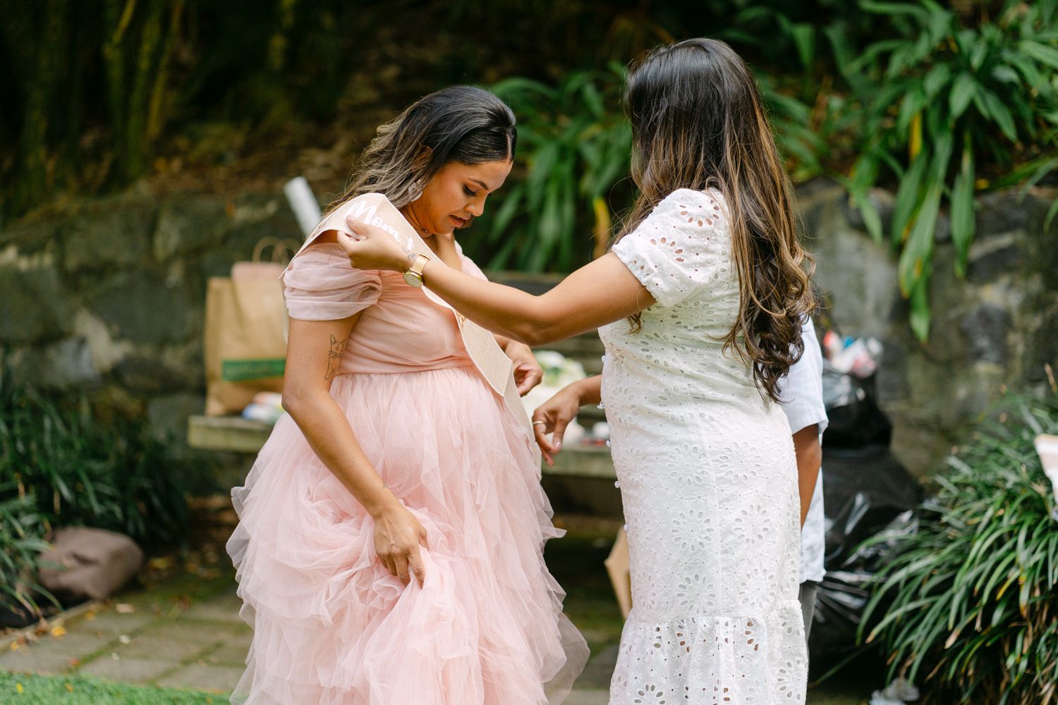 Two women in elegant dresses, one pink tulle and one white lace, interact outdoors at a garden wedding celebration.