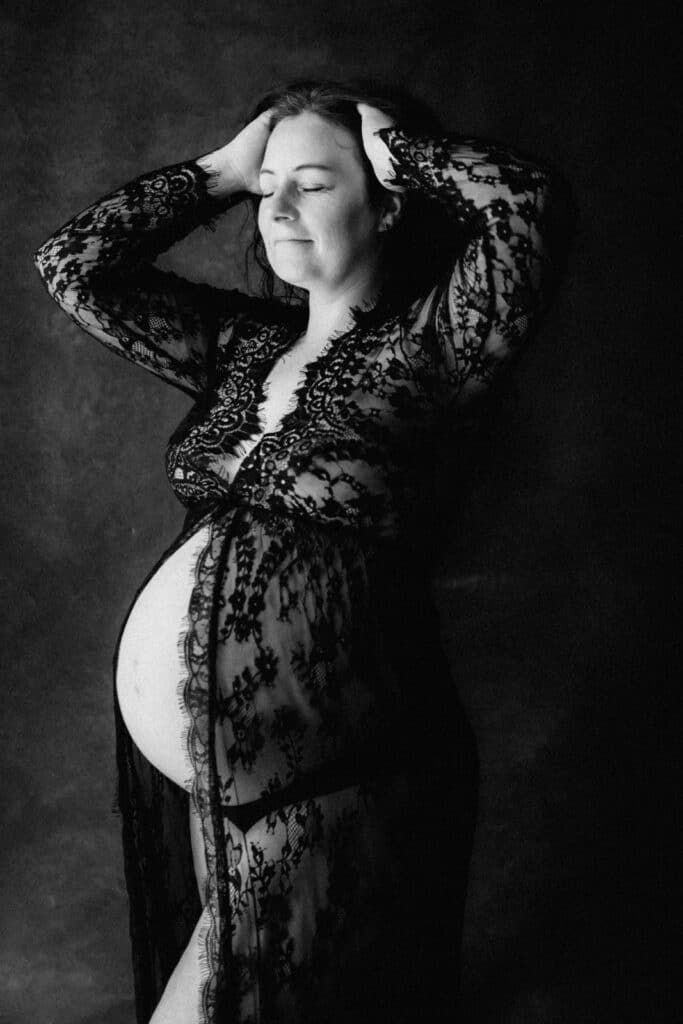 Black and white maternity photo of pregnant woman in lace robe, hands in hair, eyes closed, elegant studio portrait.