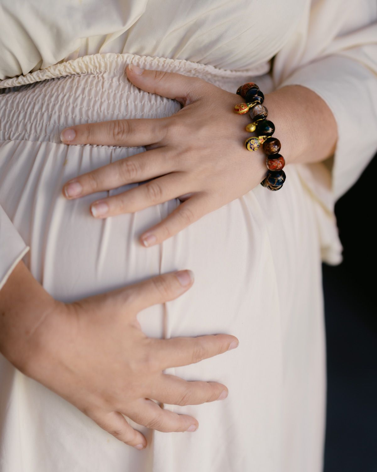 Pregnant woman in white dress cradling baby bump, wearing a dark beaded bracelet on her wrist.