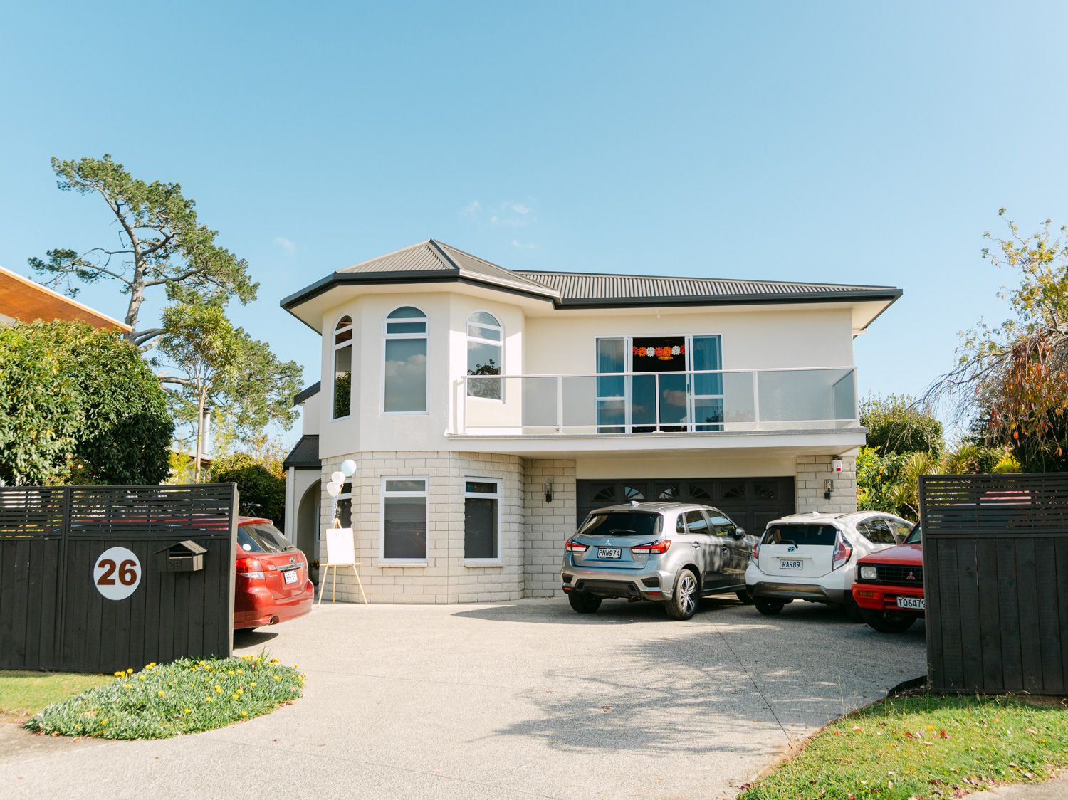Two-story modern home with balcony, large driveway with multiple cars parked, and lush trees under a clear blue sky.